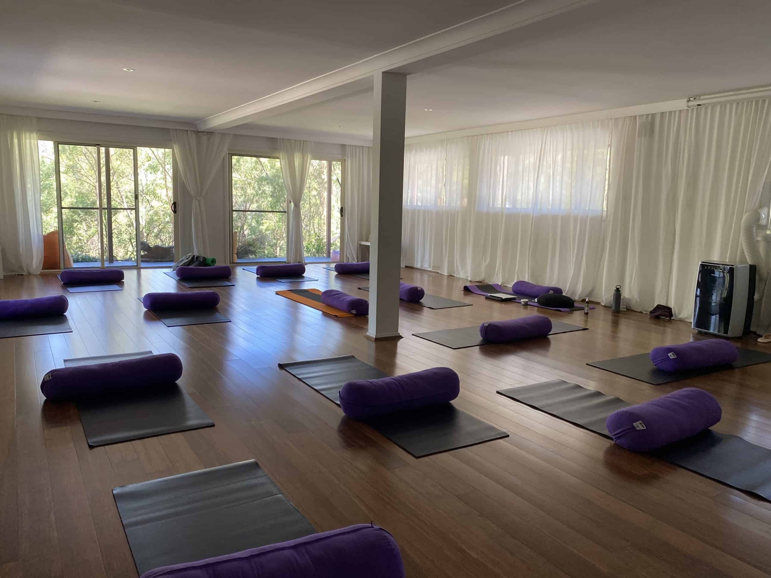 Interior of the serene studio at Yanada Retreat, NSW, prepared for a Mystique Moksha Samatvam weekend; rows of purple yoga bolsters and black mats arranged on a polished wooden floor, brightened by natural light through large windows looking out to a forest.
