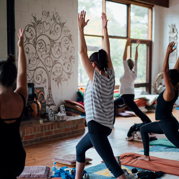 A low-angle photo from the rear of several women holding a Warrior II (Virabhadrasana) pose in a rustic, cozy studio at Mystique Moksha. The room features a wooden floor, a brick fireplace with a decorative Om tree mural, and large windows looking out onto green trees.