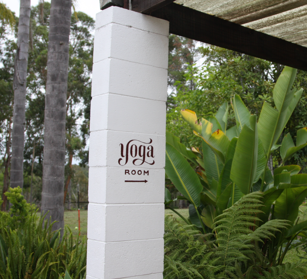 A clean white painted concrete pillar in a lush tropical garden features a dark brown sign that reads 'yoga ROOM' with an arrow pointing to the right.